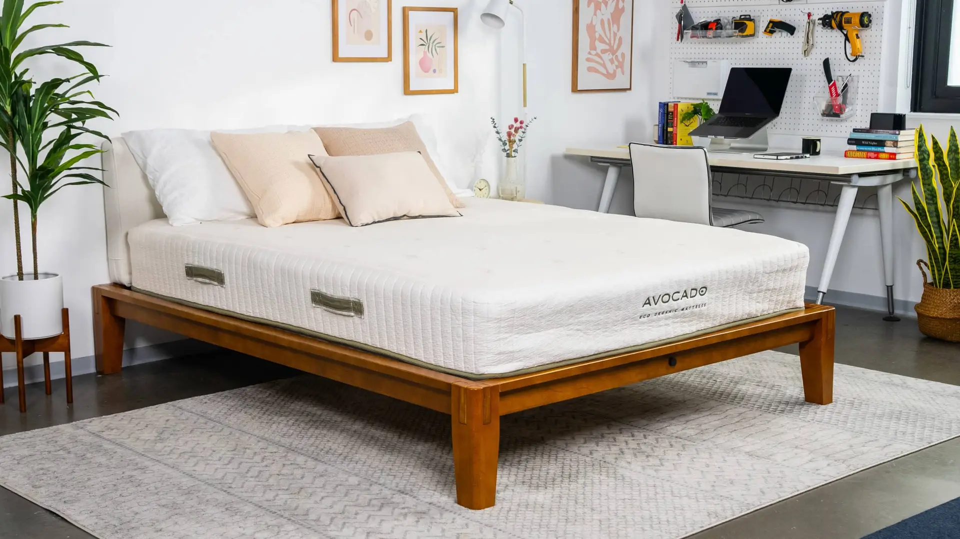 Avocado Eco Organic Mattress on wooden bed frame in modern bedroom setting with decorative pillows and workspace in background.
