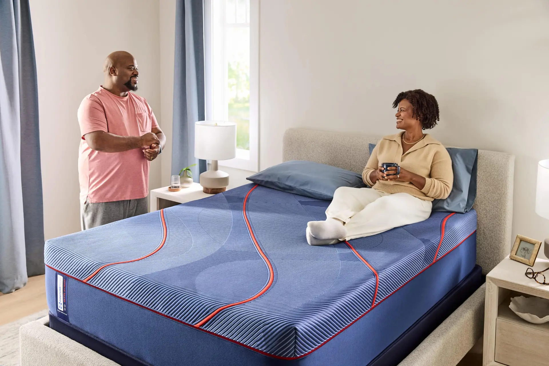 Sealy Brenham Ii Euro Pillow Top 3 Man and woman interacting in a bedroom, showcasing the Sealy Brenham II Euro Pillow Top mattress with blue and red design elements, emphasizing comfort and support.