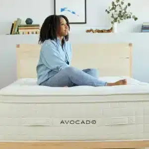 Woman sitting on Avocado Luxury Organic Pillow Top mattress, showcasing comfort and design in a modern bedroom setting.