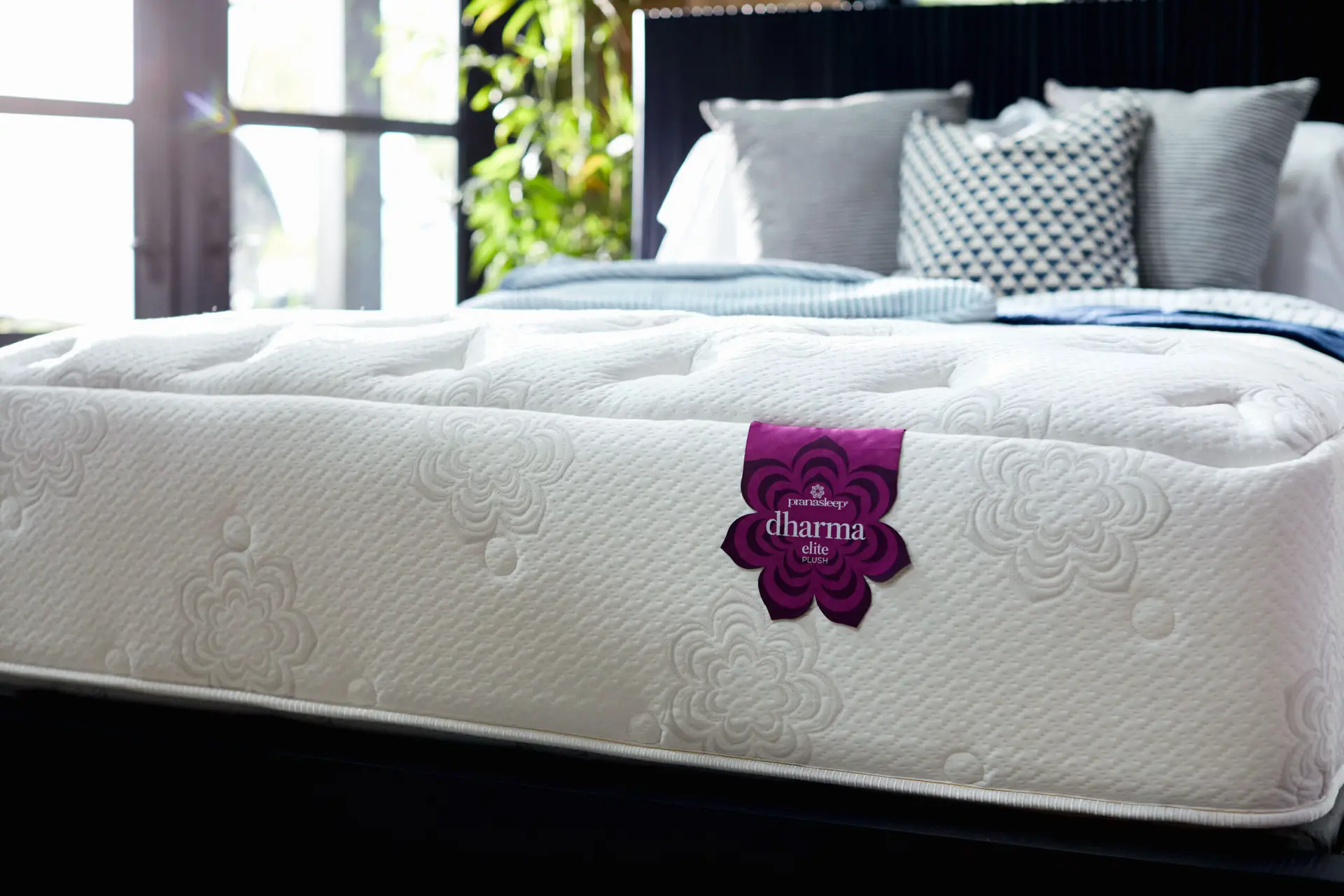 Pranasleep Dharma Elite Hybrid 2 PranaSleep Dharma Elite Hybrid mattress with natural Talalay latex, featuring a floral design, luxurious quilting, and a prominent purple "dharma" label, set against a backdrop of decorative pillows and greenery.