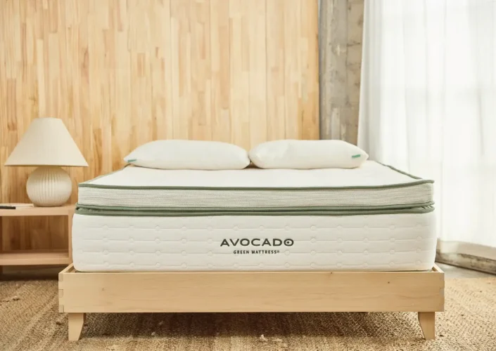 Avocado Green Mattress on a wooden bed frame, featuring organic materials and a minimalist design, with decorative pillows and a lamp in a bright, airy room.