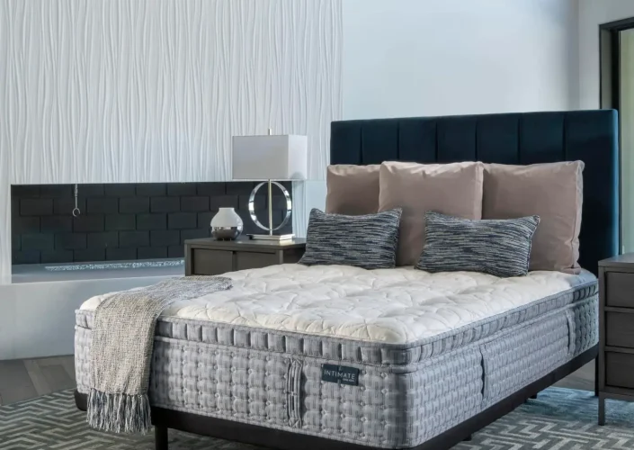 King Koil Huntington Tight Top Plush mattress on a stylish bed frame with decorative pillows, set in a modern bedroom environment featuring a textured wall and bedside lamp.