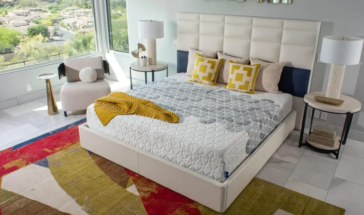 Modern bedroom featuring the King Koil Huntington Tight Top Plush mattress with a stylish quilted cover, decorative pillows, and a cozy mustard throw, set against a bright window view and contemporary decor.